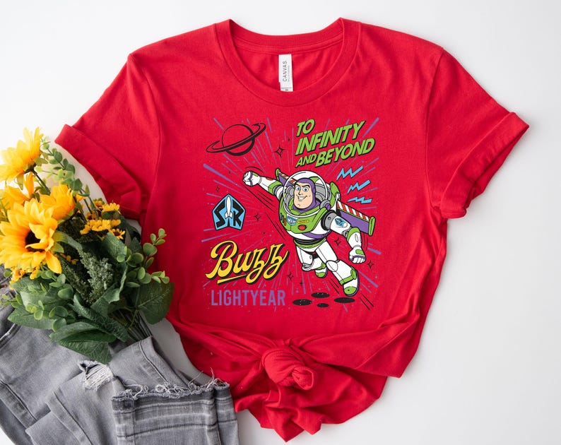 May include: A bright red short-sleeved t-shirt featuring a graphic of Buzz Lightyear in flight. The design includes the text TO INFINITY AND BEYOND, Buzz, and LIGHTYEAR, along with a planet, stars, and a space ranger emblem in blue, green, and yellow.