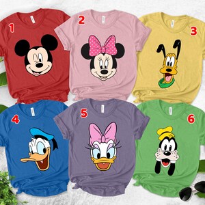 May include: A collection of six short-sleeved t-shirts featuring Disney characters. The shirts are in various colors, including red, pink, yellow, blue, purple, and green. Each shirt displays a different character: Mickey Mouse, Minnie Mouse, Pluto, Donald Duck, Daisy Duck, and Goofy.