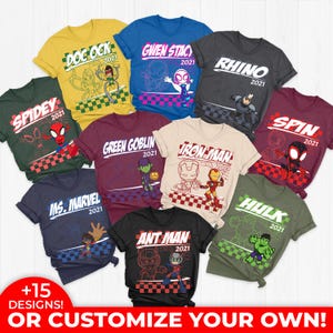 May include: A collection of colorful t-shirts featuring various superhero characters and logos. Each shirt displays a character name and the year "2021". The shirts are in various colors, including green, yellow, blue, and black, with graphic designs.