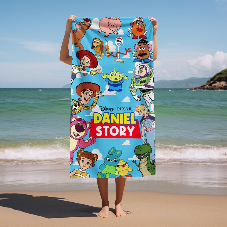 May include: A light blue rectangular beach towel featuring a colorful collage of various animated characters from the Toy Story franchise, including Woody, Buzz Lightyear, and Jessie, with the text DANIEL STORY printed in yellow and white letters.