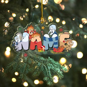 May include: A clear acrylic Christmas ornament with the word "NAME" in colorful cartoon letters. The letters are decorated with festive characters and holiday elements, including a sleigh, gifts, and snowflakes. A gold string is attached for hanging.