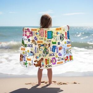 May include: A beach towel featuring various Toy Story characters and the name "JACK" repeated throughout. The towel has a light yellow background with colorful illustrations of Woody, Buzz Lightyear, and other characters. The towel is held up by a person on a sandy beach.