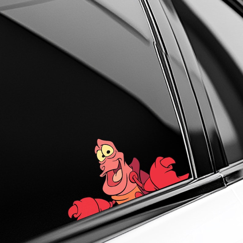 May include: A colorful sticker featuring the animated character Sebastian the crab from The Little Mermaid. The sticker shows Sebastian with a smiling expression, red claws, and a pink body. The sticker is designed to be placed on a car window.