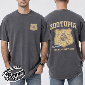 Puede incluir: Camiseta gris oscuro con un dise&ntilde;o desgastado de la insignia "Zootopia Police" en la parte delantera y trasera. La parte trasera de la camiseta muestra "Zootopia Police Department" y "Non Recruiting!" en dorado y azul.