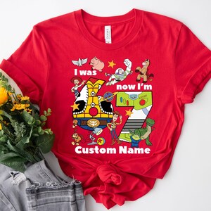 May include: Red t-shirt featuring a colorful graphic of characters from a popular animated movie. The design includes the text "I was now I'm 7 Custom Name". The shirt is a crew neck and is made of a soft material.