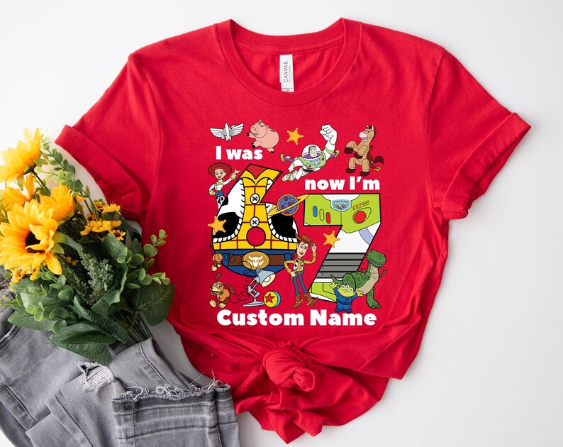 May include: Red t-shirt featuring a colorful graphic of characters from a popular animated movie. The design includes the text "I was now I'm 7 Custom Name". The shirt is a crew neck and is made of a soft material.