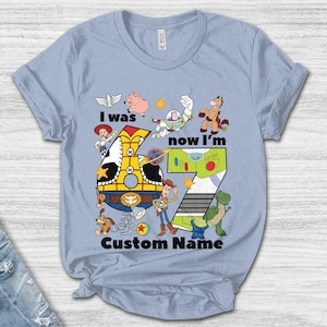 May include: Light blue t-shirt featuring a colorful graphic with the text "I was now I'm 7 Custom Name". The design includes cartoon characters and a large number 7. The shirt is made of a soft material.