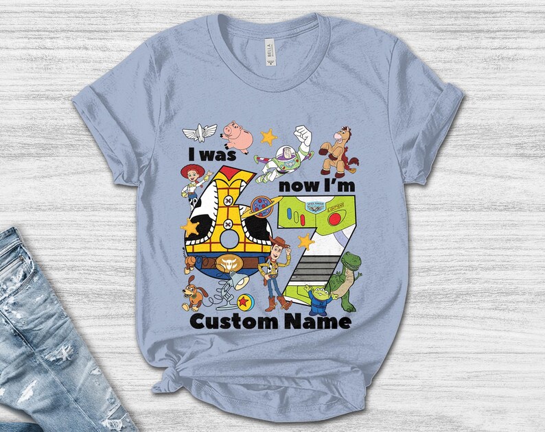 May include: Light blue t-shirt featuring a colorful graphic with the text "I was now I'm 7 Custom Name". The design includes cartoon characters and a large number 7. The shirt is made of a soft material.
