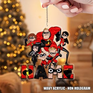 May include: A clear acrylic ornament featuring a cartoon image of a superhero family in red and black costumes. The word "NAME" is displayed in large letters at the bottom. The text "WAVY ACRYLIC - NON HOLOGRAM" is also visible.