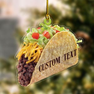 May include: A clear, taco-shaped ornament with a golden-brown shell, filled with ground meat, shredded cheese, lettuce, and diced tomatoes. The text "CUSTOM TEXT" is printed on the shell. A gold string is attached for hanging.