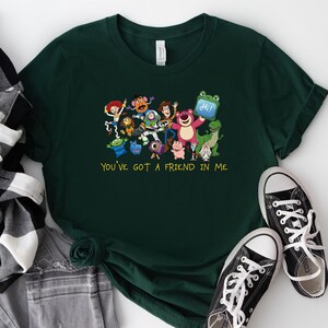 May include: A dark green short-sleeved t-shirt featuring a colorful graphic of various animated toy characters and the yellow text "YOU'VE GOT A FRIEND IN ME" printed below the group. The shirt has a crew neckline and a standard fit design.