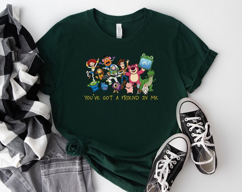 May include: A dark green short-sleeved t-shirt featuring a colorful graphic of various animated toy characters and the yellow text "YOU'VE GOT A FRIEND IN ME" printed below the group. The shirt has a crew neckline and a standard fit design.