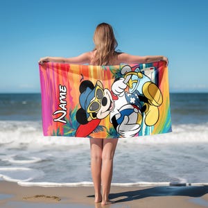 May include: A rectangular beach towel featuring a colorful sunset background with palm trees and a graphic of Mickey Mouse wearing sunglasses and a blue hat, holding a beach ball and a bucket. The word NAME is printed in white on the left side.