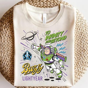 May include: A cream-colored t-shirt featuring a graphic of Buzz Lightyear in a flying pose. The design includes the text "TO INFINITY AND BEYOND" in green, "BUZZ" in yellow script, and "LIGHTYEAR" in purple, along with a planet and star icons.