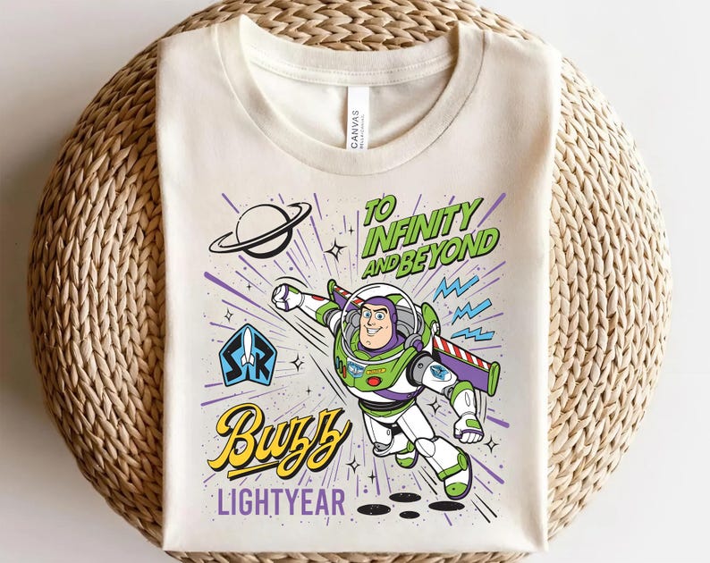 May include: A cream-colored t-shirt featuring a graphic of Buzz Lightyear in a flying pose. The design includes the text "TO INFINITY AND BEYOND" in green, "BUZZ" in yellow script, and "LIGHTYEAR" in purple, along with a planet and star icons.
