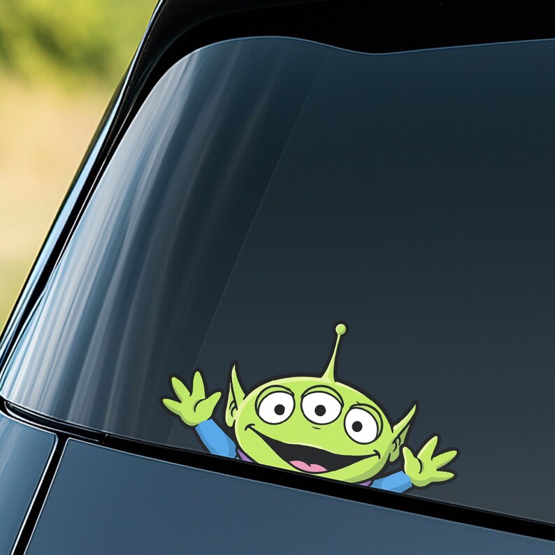 May include: A green alien character sticker with three eyes, a smiling mouth, and raised arms. The sticker is applied to a car window, with the alien's head and arms visible. The alien is wearing a blue shirt.