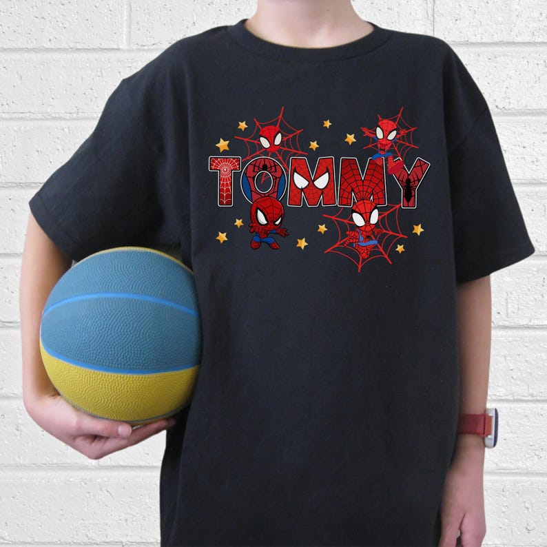 Puede incluir: Camiseta negra con el nombre "TOMMY" en rojo y azul, con gr&aacute;ficos de Spider-Man y estrellas amarillas. La camiseta tiene mangas cortas. Una pelota de baloncesto azul y amarilla se sostiene en la mano.