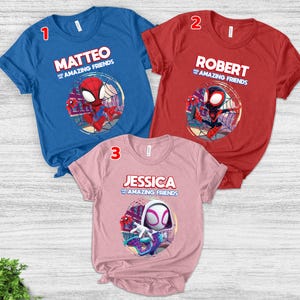 Op de afbeelding: Drie shirts met korte mouwen in blauw, rood en roze. Elk shirt heeft een ander Spider-Man-personageontwerp met een naam en de zin "and the Amazing Friends". De shirts worden gepresenteerd op een licht houten oppervlak.