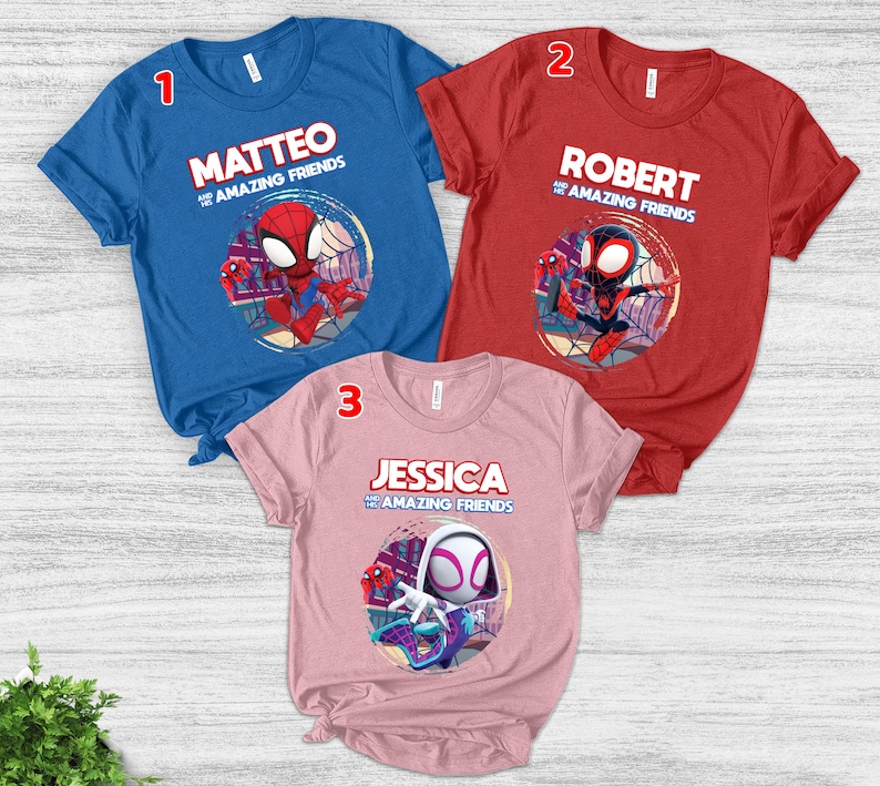 Op de afbeelding: Drie shirts met korte mouwen in blauw, rood en roze. Elk shirt heeft een ander Spider-Man-personageontwerp met een naam en de zin "and the Amazing Friends". De shirts worden gepresenteerd op een licht houten oppervlak.