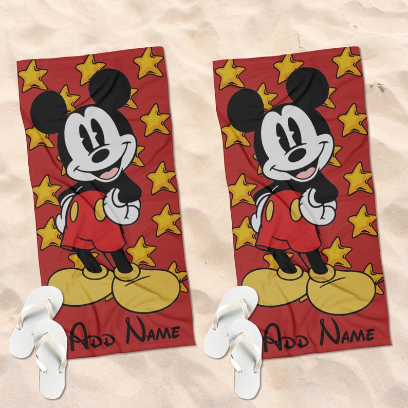 May include: Two rectangular beach towels featuring a red background with yellow stars and a graphic of Mickey Mouse wearing red shorts and yellow shoes. Each towel includes the black text Add Name at the bottom, accompanied by white flip-flops.