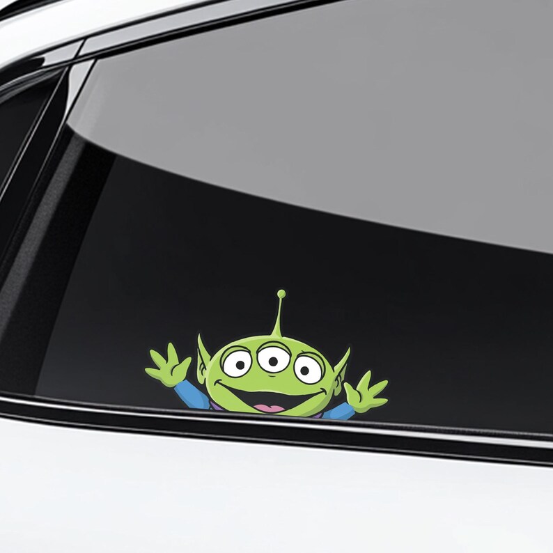 May include: A green alien character sticker with three eyes, large ears, and a smiling expression. The sticker is applied to a car window, with the alien's arms raised in a welcoming gesture. The design is a cartoon style.