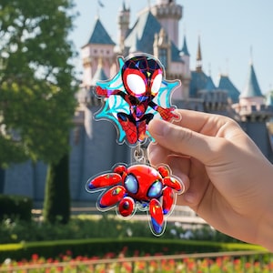 May include: A Spider-Man keychain featuring two acrylic charms. The top charm is a cartoon Spider-Man figure with a blue web design. The bottom charm is a red and blue spider. The background includes a castle and greenery.