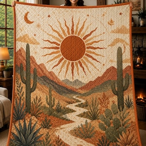 May include: A rectangular quilt featuring a desert landscape with a large central sun, a winding path, saguaro cacti, and agave plants. The design includes a crescent moon and stars against a cream background with orange trim and stitching.