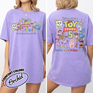 May include: A lilac-colored t-shirt featuring the Toy Story 5 logo and characters. The front of the shirt displays the logo and characters, while the back has a larger design with the text "You've Got a Friend In Me."