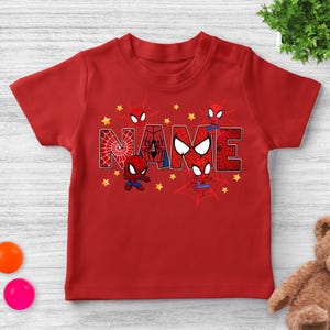 Puede incluir: Camiseta roja con la palabra "NAME" en un estilo de letras en bloque, con un tema de Spider-Man. Las letras est&aacute;n decoradas con telara&ntilde;as y personajes de Spider-Man. Estrellas amarillas est&aacute;n dispersas alrededor de las letras.