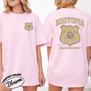 Puede incluir: Camiseta rosa con un dise&ntilde;o de insignia de polic&iacute;a dorada y marr&oacute;n en la parte delantera y trasera. La parte trasera de la camiseta presenta la palabra "ZOOTOPIA" encima de la insignia y "POLICE DEPARTMENT" debajo. La camiseta est&aacute; etiquetada como "Comfort Colors Blossom".
