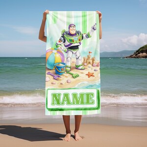 May include: A rectangular beach towel featuring a cartoon space ranger character standing on a sandy beach with a beach ball, a blue bucket, a sandcastle, and a starfish. The towel has a green and white striped background and a green box with the text NAME.
