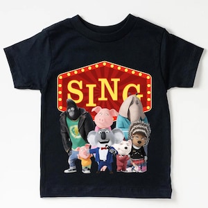 May include: Black t-shirt featuring the word "SING" in yellow letters within a red marquee design. The shirt showcases a group of animated characters from the movie "Sing", including a gorilla, pig, elephant, and koala.