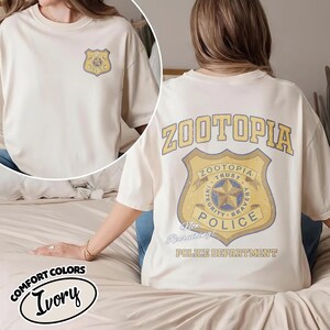 Puede incluir: Camiseta color marfil con un dise&ntilde;o de insignia del Departamento de Polic&iacute;a de Zootopia en la parte delantera y trasera. La parte trasera de la camiseta muestra la palabra "Zootopia" encima de la insignia, con el texto "Not Recruiting!" debajo.
