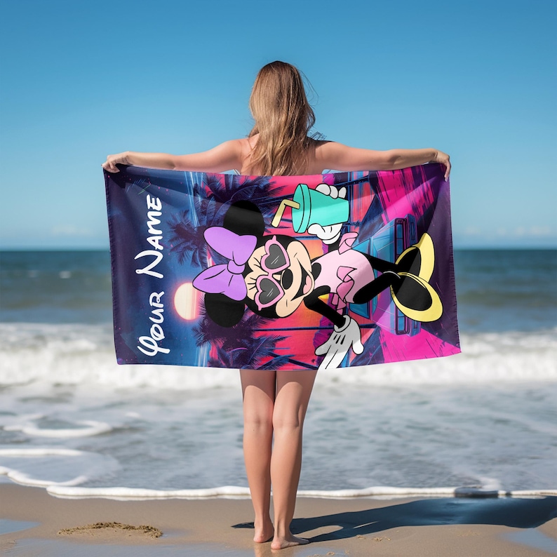Op de afbeelding: Een rechthoekige strandhanddoek met een afbeelding van een tekenfilmmuis met een hartvormige zonnebril en een paarse strik, die een turquoise drankje vasthoudt. De achtergrond toont een zonsondergang met palmbomen. Tekst YOUR NAME staat links.