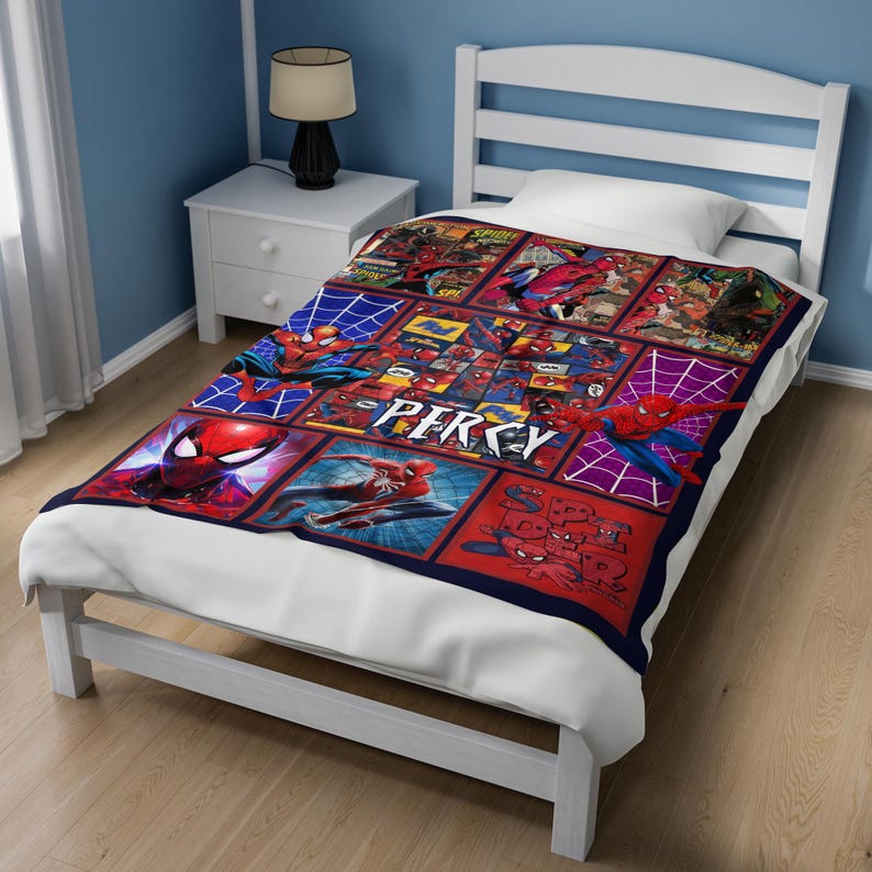 May include: A red, blue, and white Spider-Man themed blanket with a grid pattern. The blanket features various Spider-Man comic book panels and the name "PERCY" in white block letters. The blanket is on a white bed.