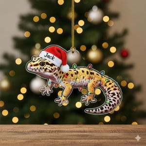 May include: A Christmas ornament featuring a leopard gecko wearing a Santa hat with the name "Jack" on it. The gecko is decorated with colorful Christmas lights. The ornament is hanging in front of a blurred Christmas tree.