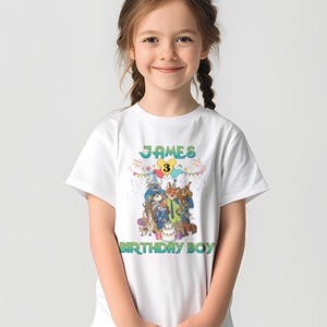 May include: A white short-sleeved t-shirt featuring a colorful graphic of various cartoon animals wearing party hats around a birthday cake. The text reads JAMES at the top, the number 3 in a yellow circle, and BIRTHDAY BOY at the bottom.