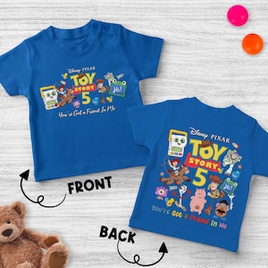 May include: Two blue Toy Story 5 t-shirts. The front shirt has the text "Toy Story 5" with cartoon characters and the phrase "You're Got a Friend In Me." The back shirt has the same design. The shirts are on a white wooden surface.