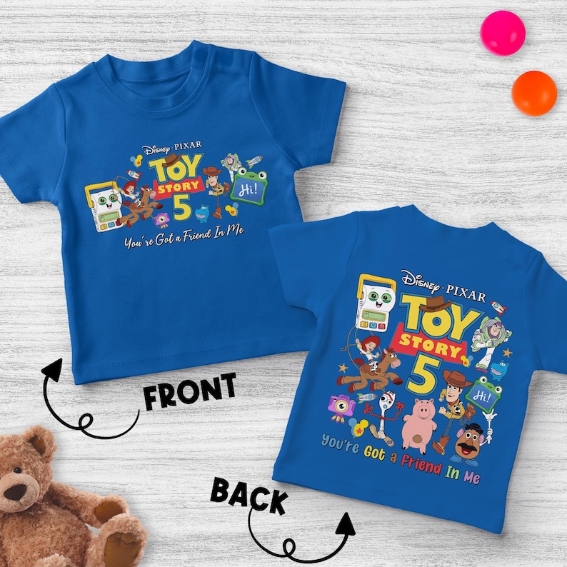 May include: Two blue Toy Story 5 t-shirts. The front shirt has the text "Toy Story 5" with cartoon characters and the phrase "You're Got a Friend In Me." The back shirt has the same design. The shirts are on a white wooden surface.
