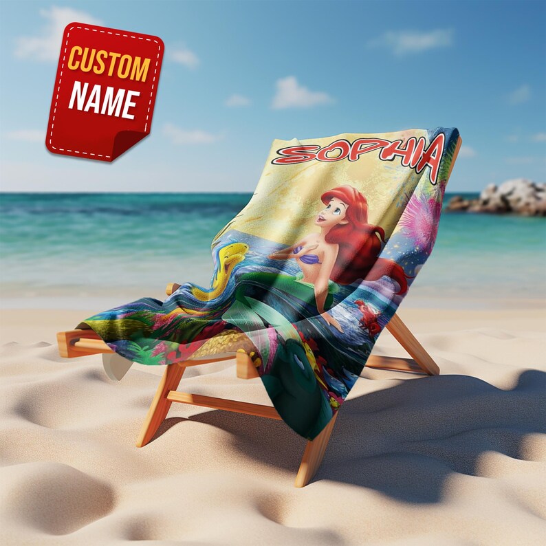 Puede incluir: Una toalla de playa sobre una silla de madera con el gr&aacute;fico de una sirena pelirroja con cola verde, un pez amarillo y un cangrejo. El nombre SOPHIA est&aacute; impreso en letras rojas grandes en la parte superior sobre un fondo colorido.