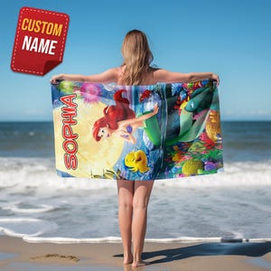 Puede incluir: Una toalla de playa rectangular con un gr&aacute;fico colorido de una sirena de pelo rojo y un pez amarillo sobre un fondo submarino a la luz de la luna. El nombre SOPHIA est&aacute; impreso en letras rojas grandes en el lado izquierdo del dise&ntilde;o.