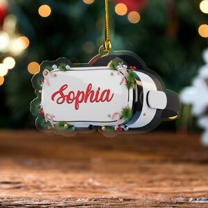 May include: A VR headset Christmas ornament with the name "Sophia" in red script. The white panel is decorated with candy canes, greenery, and silver ornaments. The headset is black and white, with a gold hanging string.