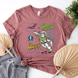 May include: A dusty rose t-shirt featuring a graphic of Buzz Lightyear in flight. The design includes the text "TO INFINITY AND BEYOND" in green, "Buzz" in yellow script, and "LIGHTYEAR" in purple, alongside a planet and star icons.