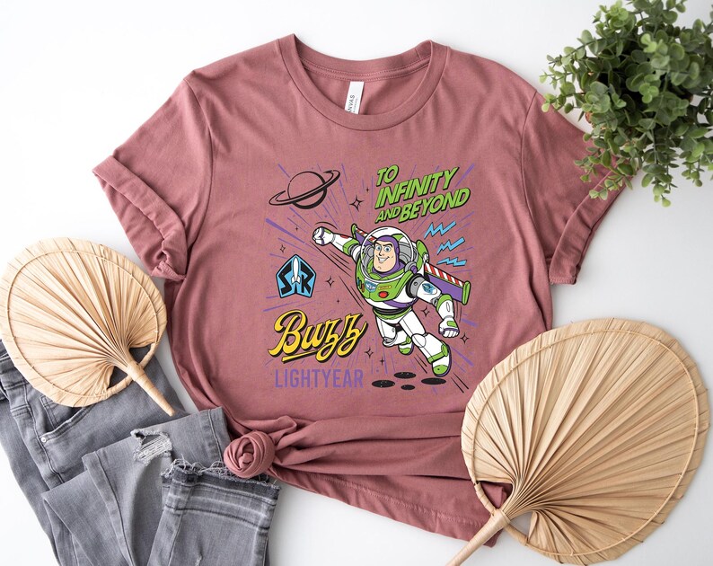 May include: A dusty rose t-shirt featuring a graphic of Buzz Lightyear in flight. The design includes the text "TO INFINITY AND BEYOND" in green, "Buzz" in yellow script, and "LIGHTYEAR" in purple, alongside a planet and star icons.