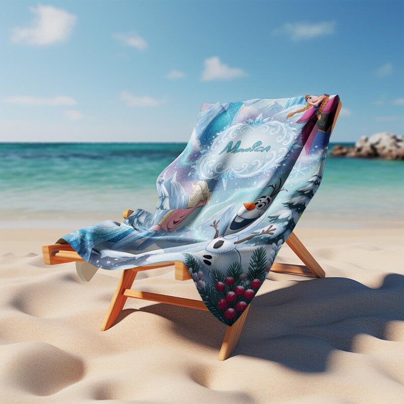May include: A beach chair on a sandy beach with a blue and white Frozen-themed towel draped over it. The towel features images of Elsa, Anna, and Olaf, with the word "Maui" in a decorative font. The background shows the ocean and sky.