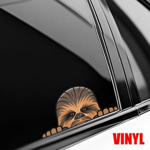 May include: A brown and tan vinyl decal featuring a cartoon image of Chewbacca peeking over a car window. The word "VINYL" is printed in red at the bottom right corner. The decal is designed to be applied to a car window.
