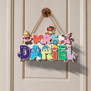 May include: A decorative wooden wall sign featuring the text "MRS. DANIEL" in colorful, bold letters. The sign is adorned with various animated character illustrations from the movie Inside Out and hangs from a brown rope on a wooden peg.