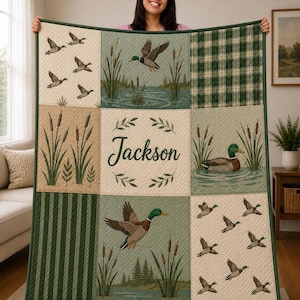 May include: A quilted blanket featuring a grid of nine panels with mallard ducks, cattails, and green plaid patterns. The center panel displays the name Jackson in cursive script surrounded by a leaf wreath, all framed by a dark green border.