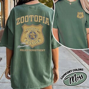 Puede incluir: Camiseta verde con dise&ntilde;o de "Zootopia Police Department". La parte trasera presenta un gran gr&aacute;fico de insignia con texto, y la parte delantera tiene una insignia m&aacute;s peque&ntilde;a. La camiseta es de color verde musgo.