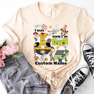 May include: A beige t-shirt featuring a colorful graphic with the text "I was now I'm" and the number 7. The graphic includes cartoon characters and the words "Custom Name". Black canvas sneakers and gray jeans are also in the image.